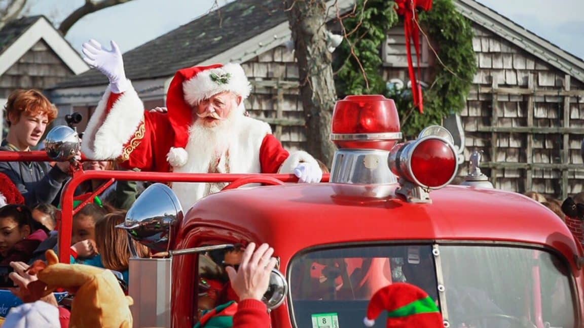 santa claus waving in a red truck with a green wreath in the background and people waving