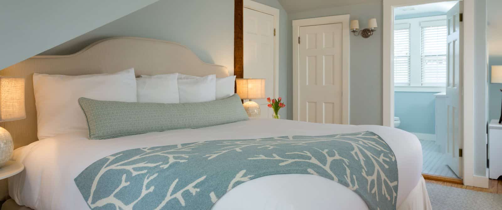 Bedroom with light blue walls, cream upholstered headboard, white bedding, and view into bathroom