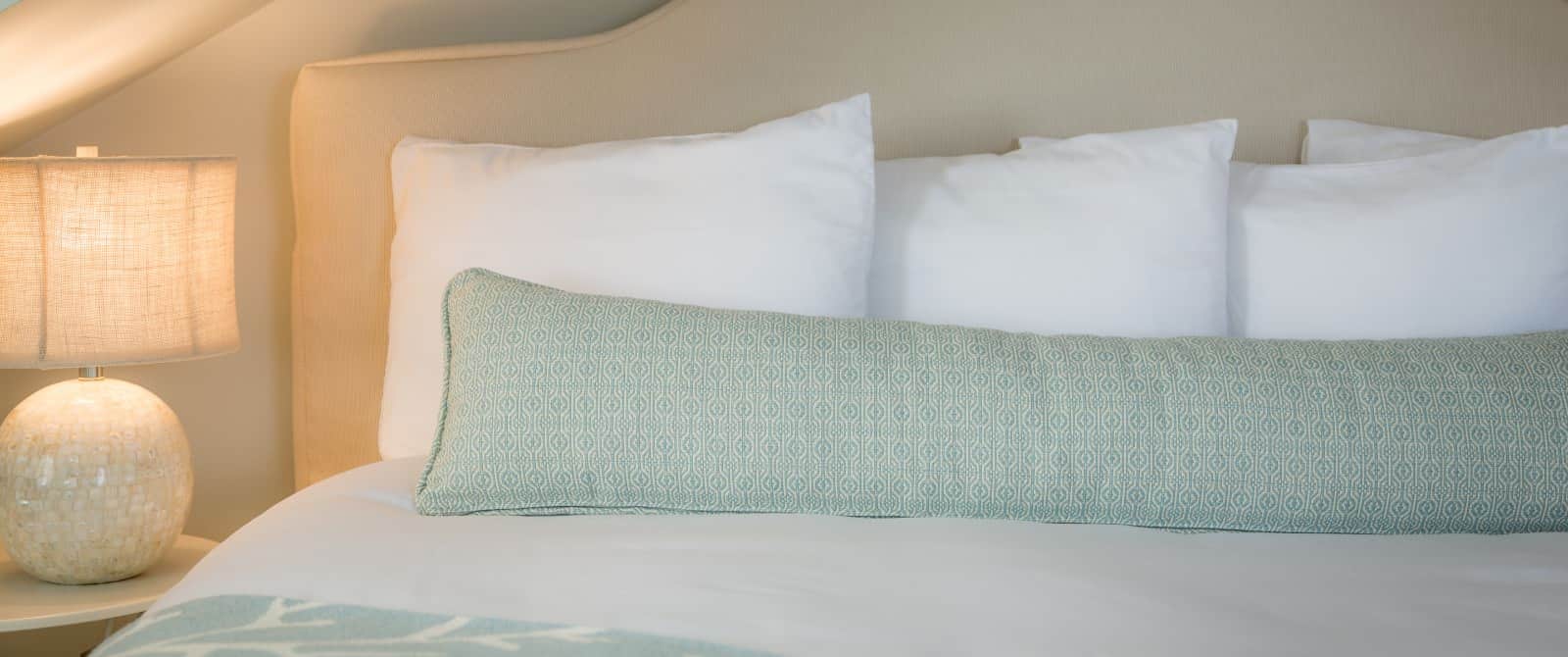 Close up view of cream upholstered headboard, white bedding, patterned light sage pillow, and lighted lamp