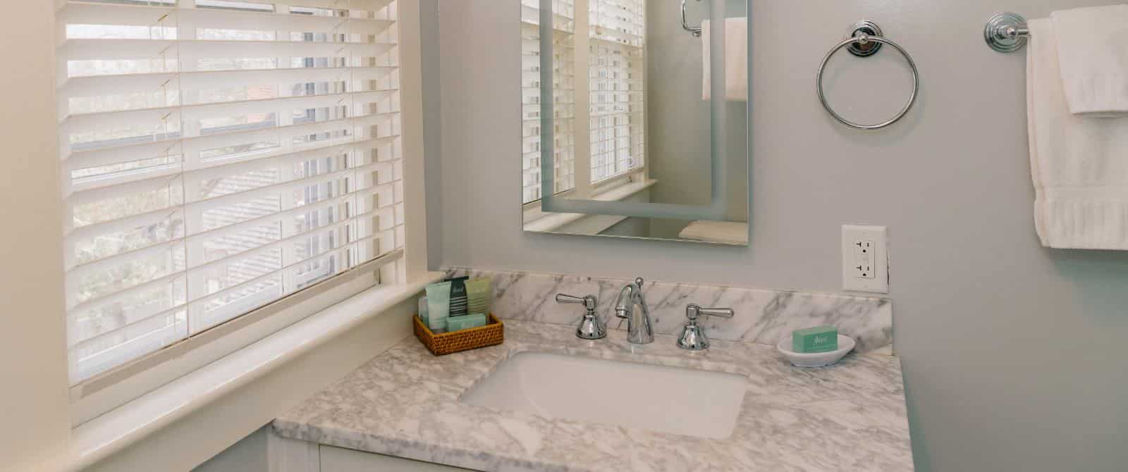 Bathroom with light colored walls, white vanity with marble top, and window with white blinds