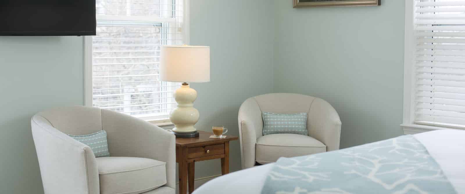 Bedroom with light green walls, white bedding, and sitting area with cream upholstered chairs