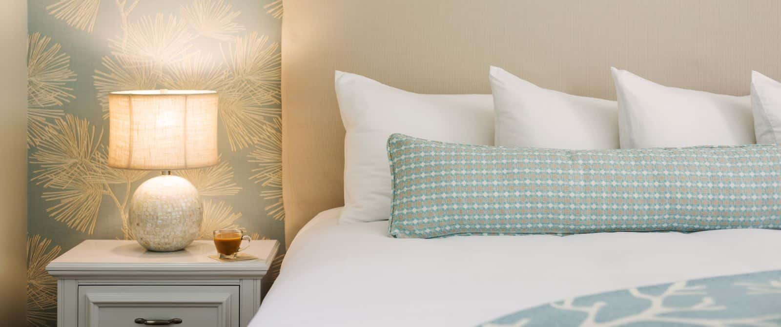 Close up view of bed with light tan upholstered headboard, white bedding, light blue and green patterned long pillow, white side table, and lighted lamp