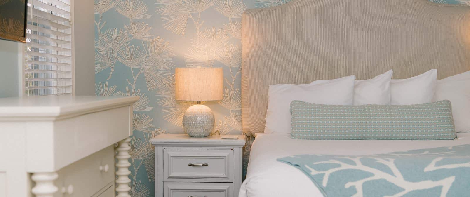 Close up view of bed with light tan upholstered headboard, white bedding, light blue and green patterned long pillow, white small dresser, and lighted lamp
