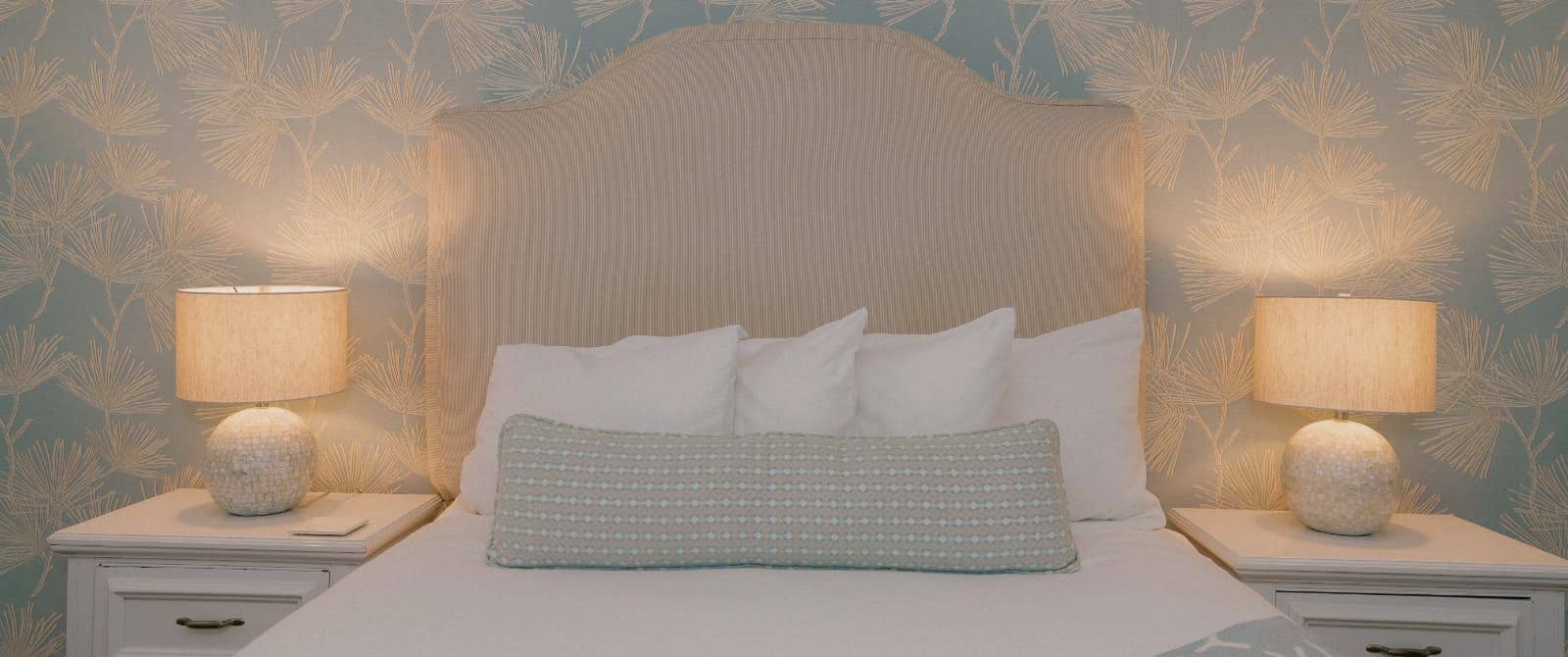 Close up view of bed with light tan upholstered headboard, white bedding, light blue and green patterned long pillow, white side tables, and lighted lamps
