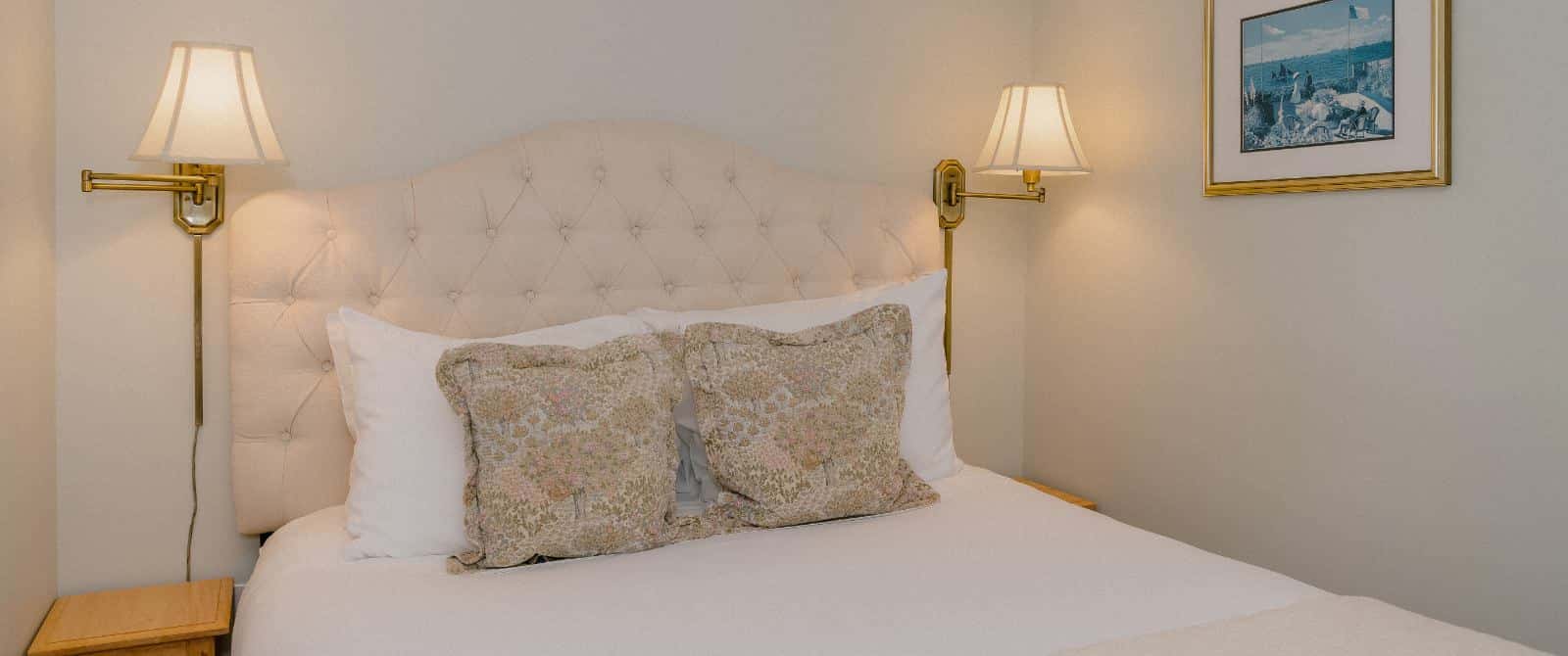 Close up view of bed with cream upholstered headboard, white bedding, and brass wall sconces