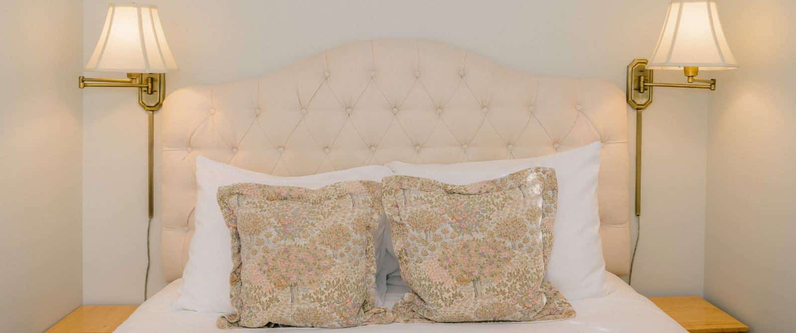 Close up view of bed with cream upholstered headboard, white bedding, and brass wall sconces