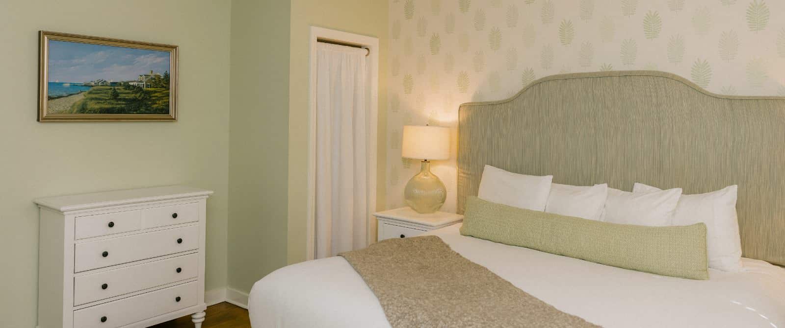 Bedroom with wooden flooring, light green walls, multicolored frawns wallpaper, light green upholstered headboard, white bedding, and white dresser