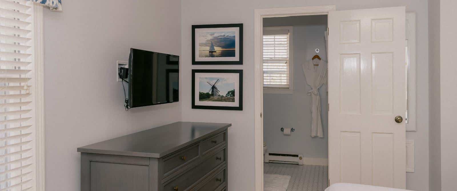 Bedroom with light colored walls, gray dresser, wall-mounted flat-screen TV, and view into bathroom