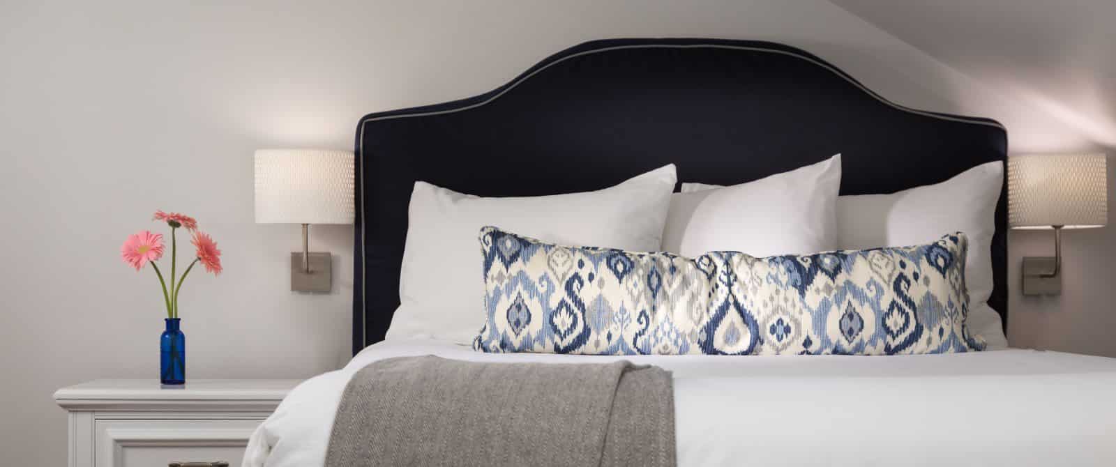Bedroom with white walls, navy upholstered headboard, white bedding, white side table, and wall-mounted sconces