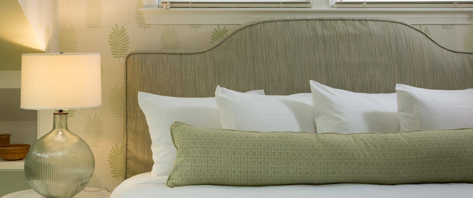 Close up view of light green upholstered headboard, white bedding, patterned light sage pillow, and lighted lamp
