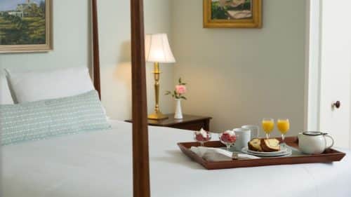 Close up view of dark wooden four-poster bed, white bedding, and dark wooden tray with fruit cups, coffee, juice, and breakfast breads