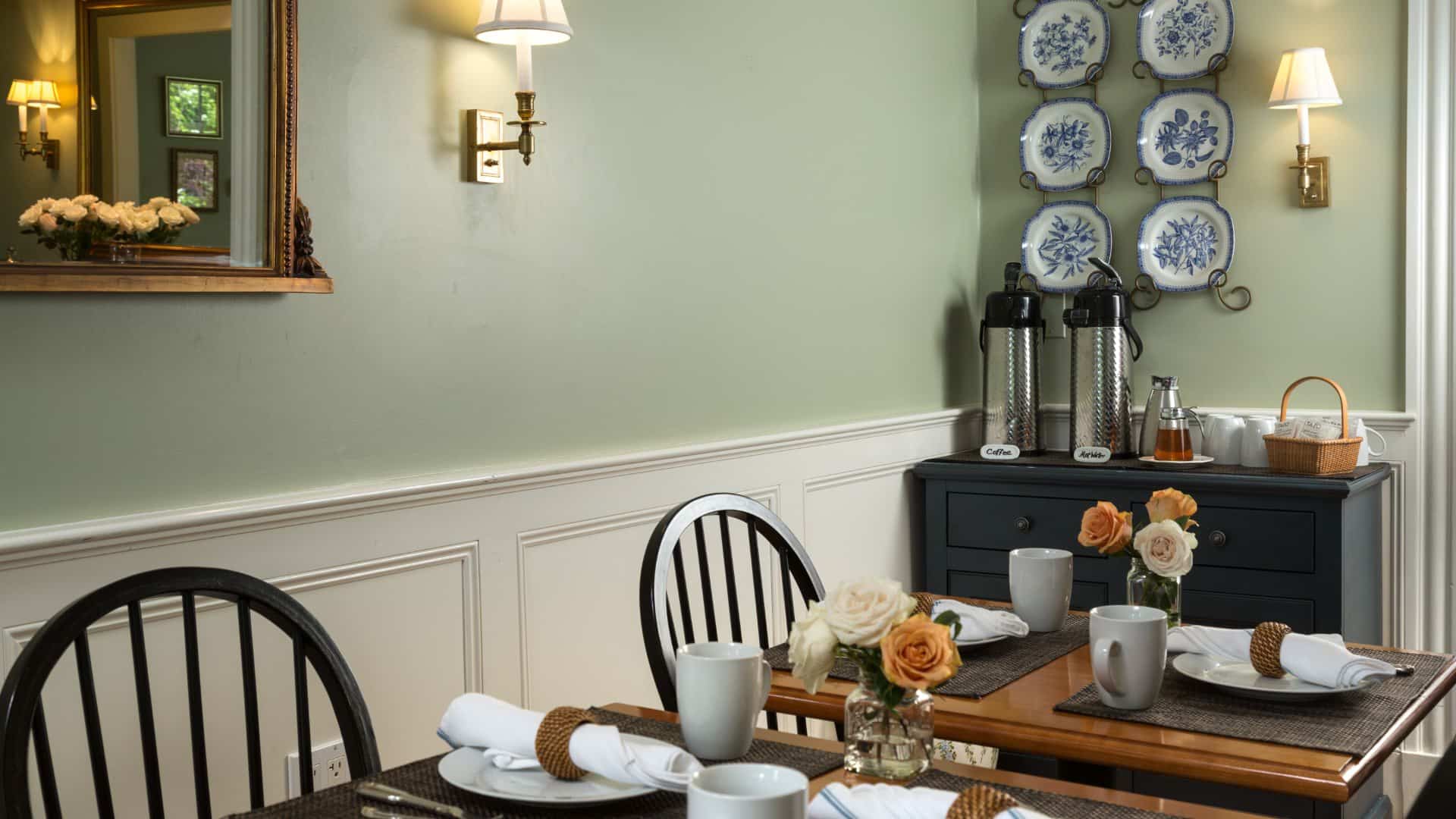 Dining area with wooden tables, black wooden chairs, light sage green walls, white wainscoting, and blue and white plates hanging on the wall