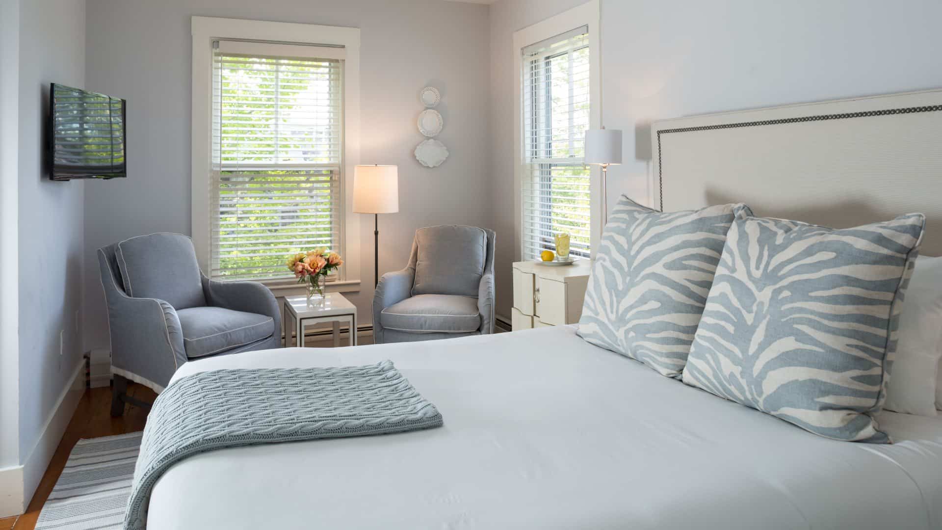 Bedroom with wooden flooring, light colored walls, white upholstered headboard, white bedding, and sitting area with two light gray upholstered chairs
