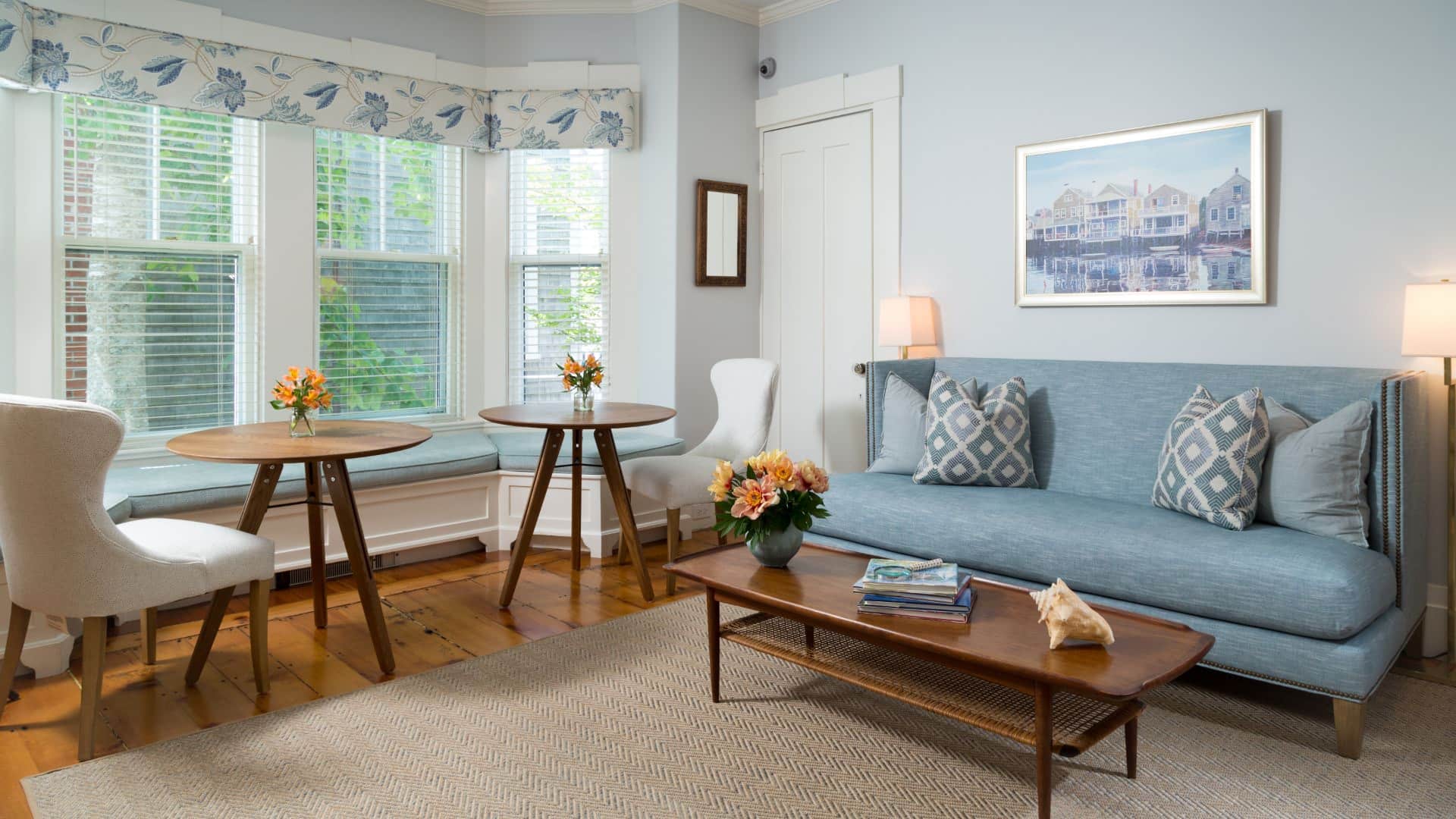 Gathering area with blue upholstered sofa, wooden coffee table, wooden flooring, light blue walls, and two small wooden tables with cream chairs