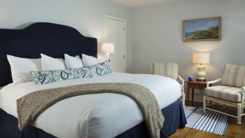 Bedroom with wooden flooring, light blue walls, navy upholstered headboard, white bedding, and sitting area with white chairs each with striped cushions