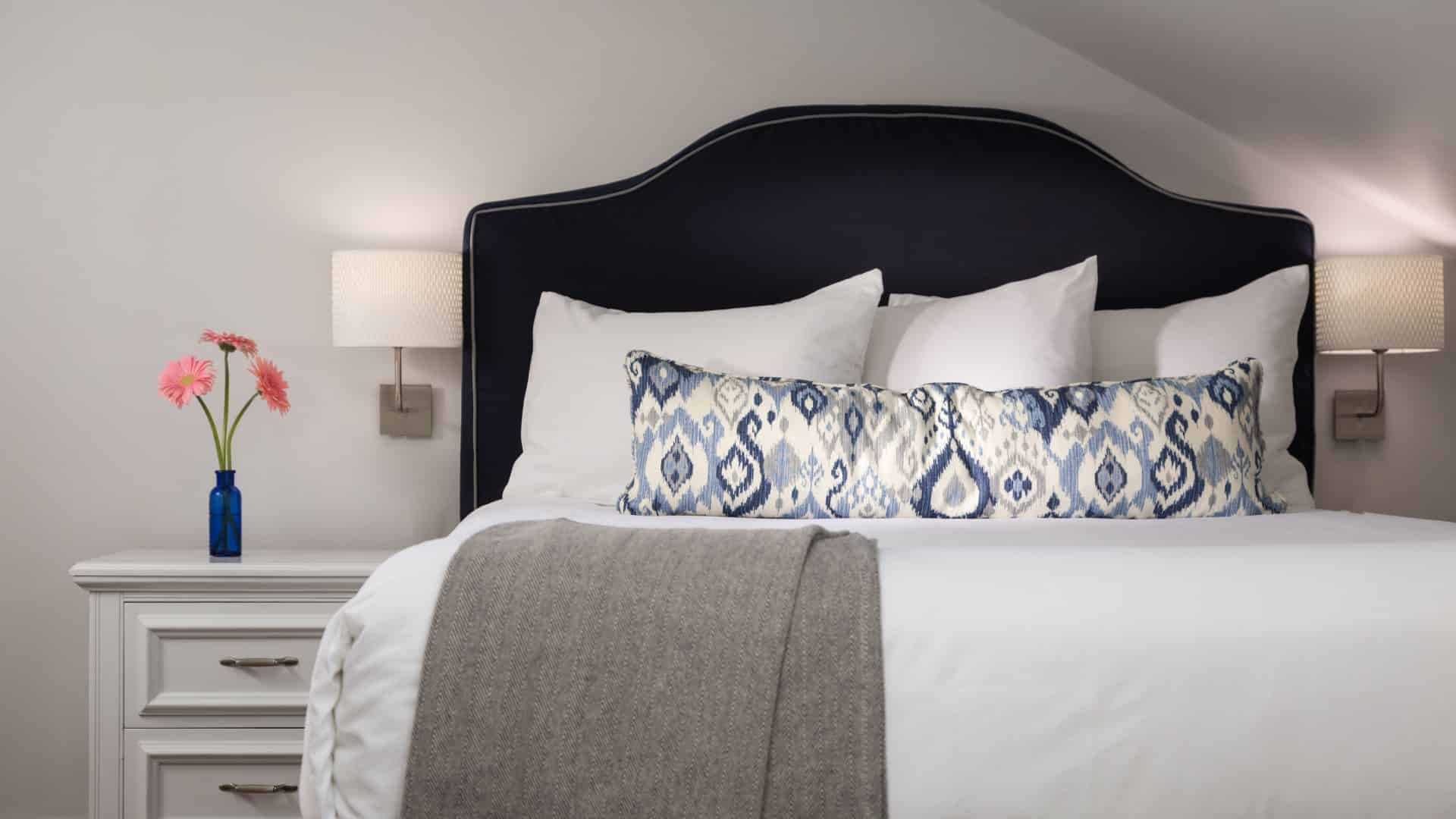 Bedroom with white walls, navy upholstered headboard, white bedding, white side table, and wall-mounted sconces