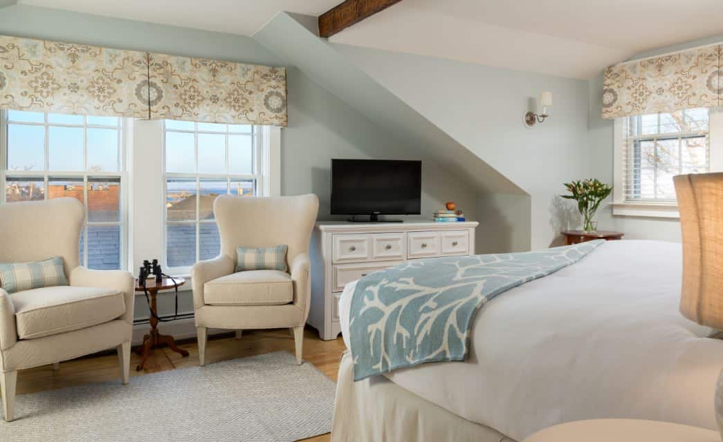 Bedroom with hardwood flooring, light blue walls, white dresser, white bedding, and cream upholstered chairs in sitting area