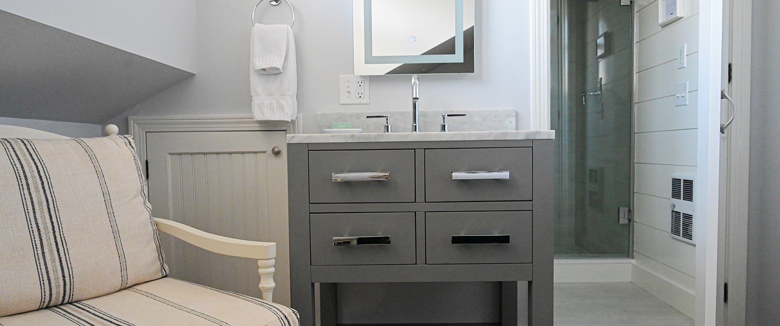 a grey vanity and a cream and blue striped chair with a door open