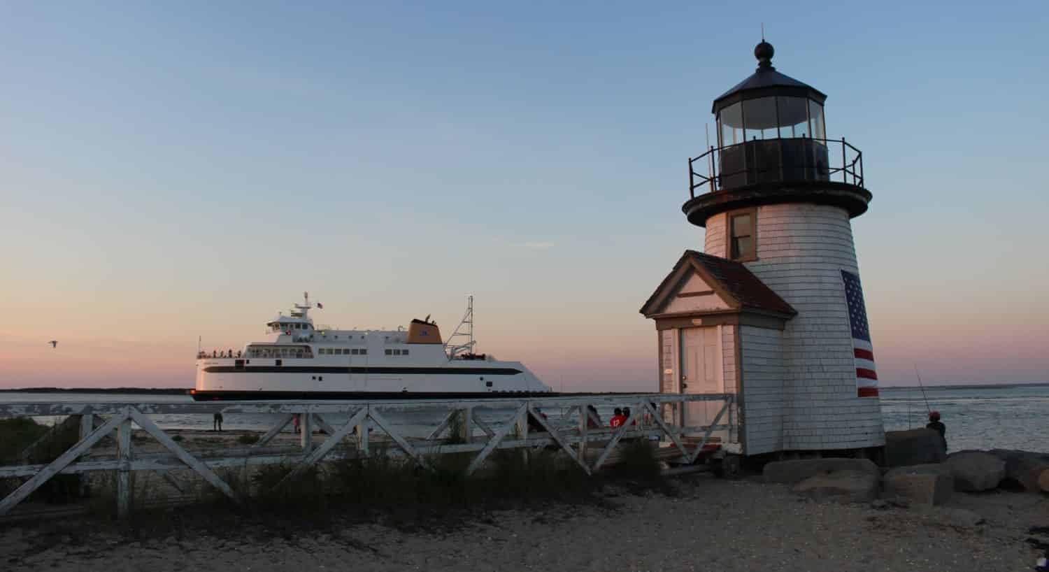 Small lighthouse next to ocean with small cruise ship in the background at dusk