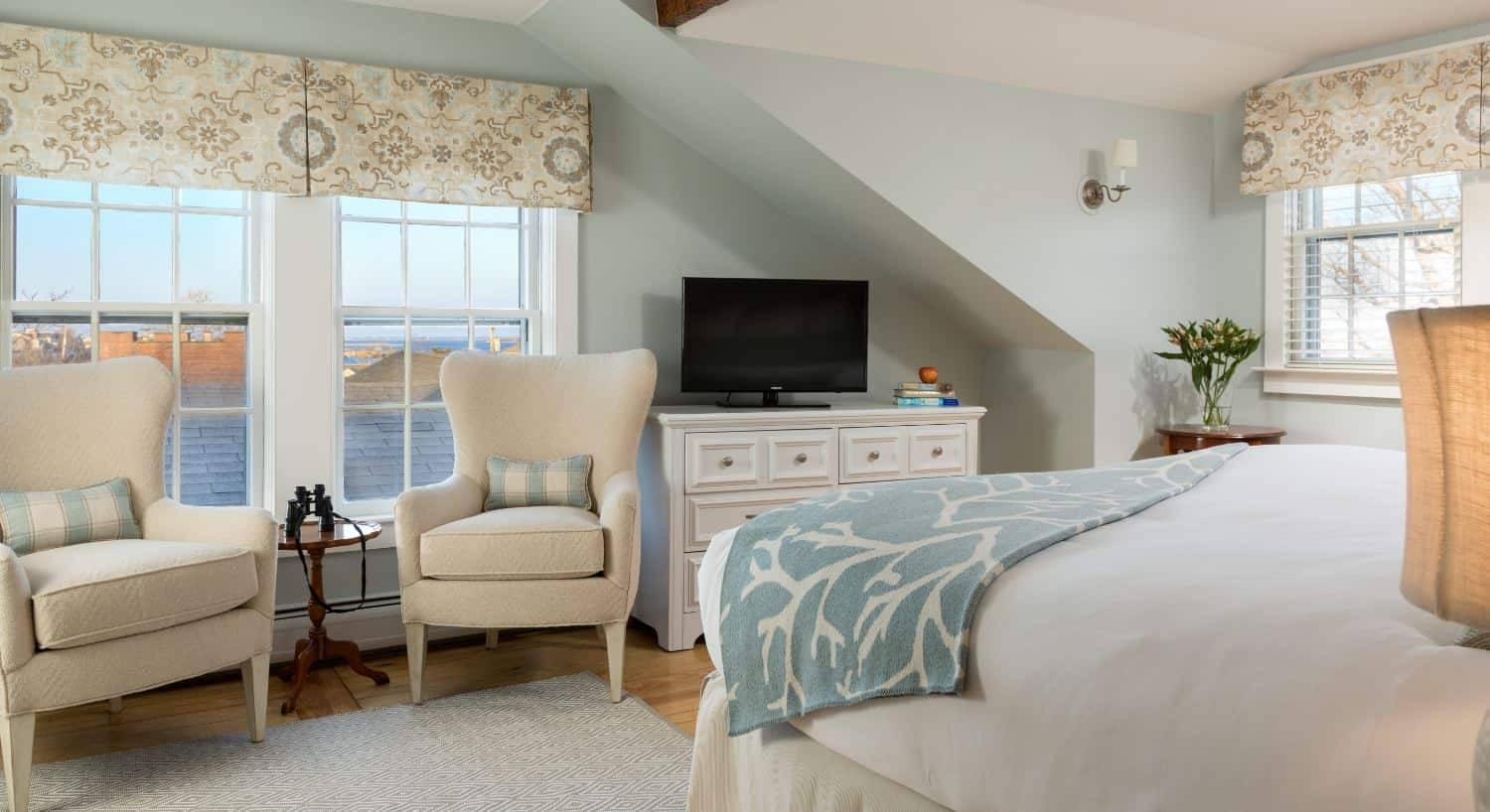 Large bedroom with wooden flooring, light blue walls, white bedding, white dresser, and sitting area with two cream upholstered chairs